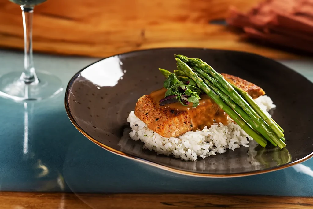Professionally plated grilled protein, rice, asparagus, brown sauce on dark plate.