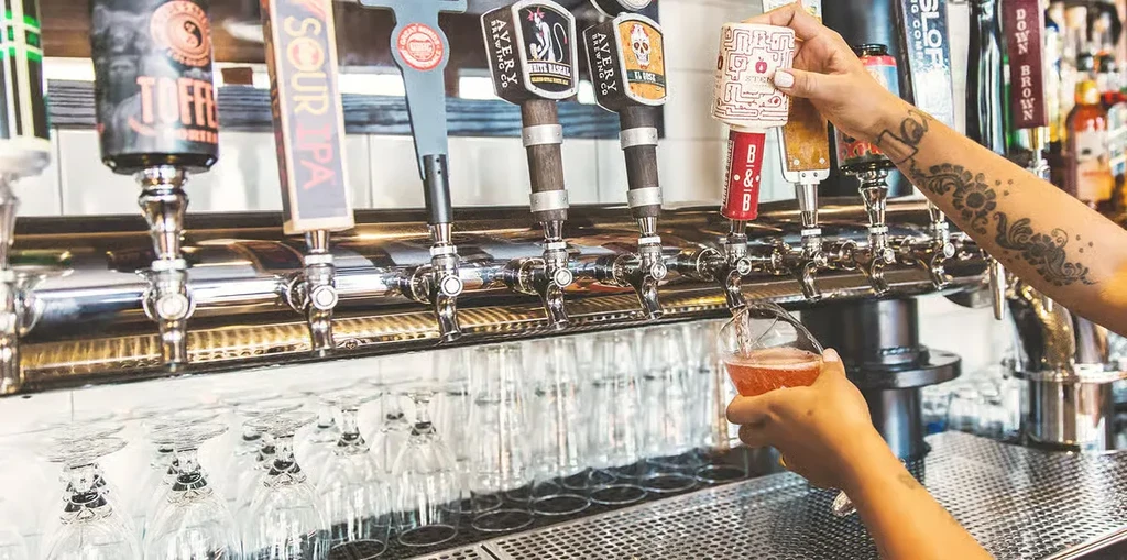 Close-up: Tattooed arm fills glass from shiny bar taps.