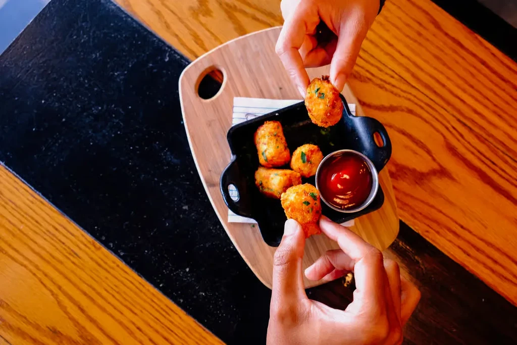 Two hands pick golden fried food from a skillet with ketchup.