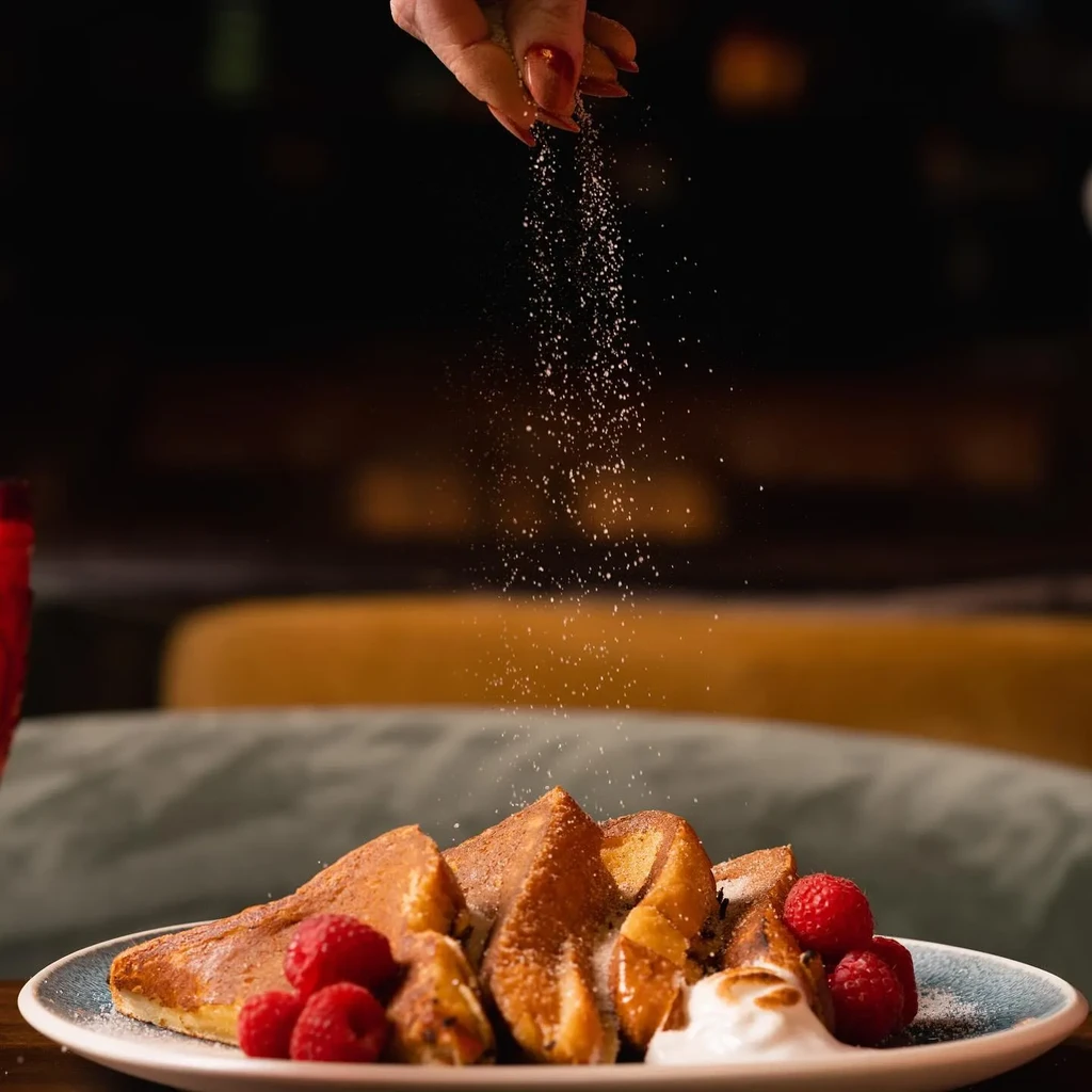 Hand sprinkles sugar on golden French toast with raspberries and cream.