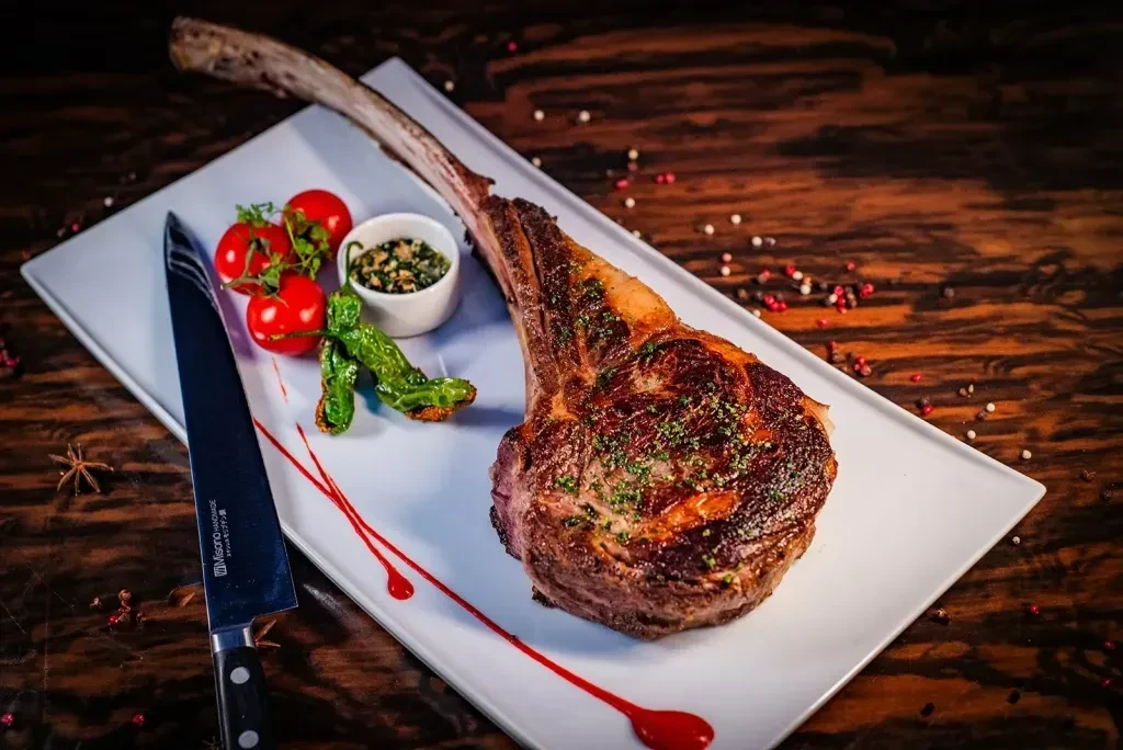 Large Tomahawk steak with tomatoes, chilis, herby sauce, and a knife.