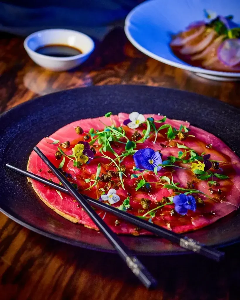 Thinly sliced tuna carpaccio with flowers, microgreens, and chopsticks.