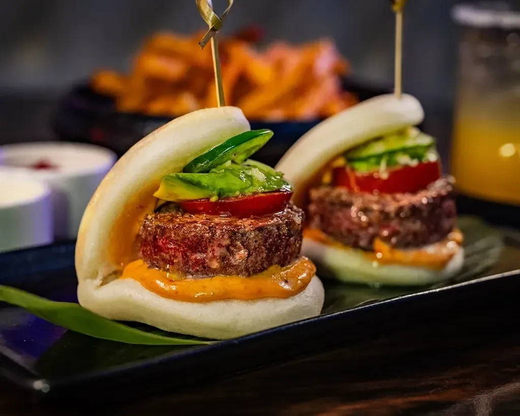 Two gourmet beef sliders on bao buns with avocado and tomato.