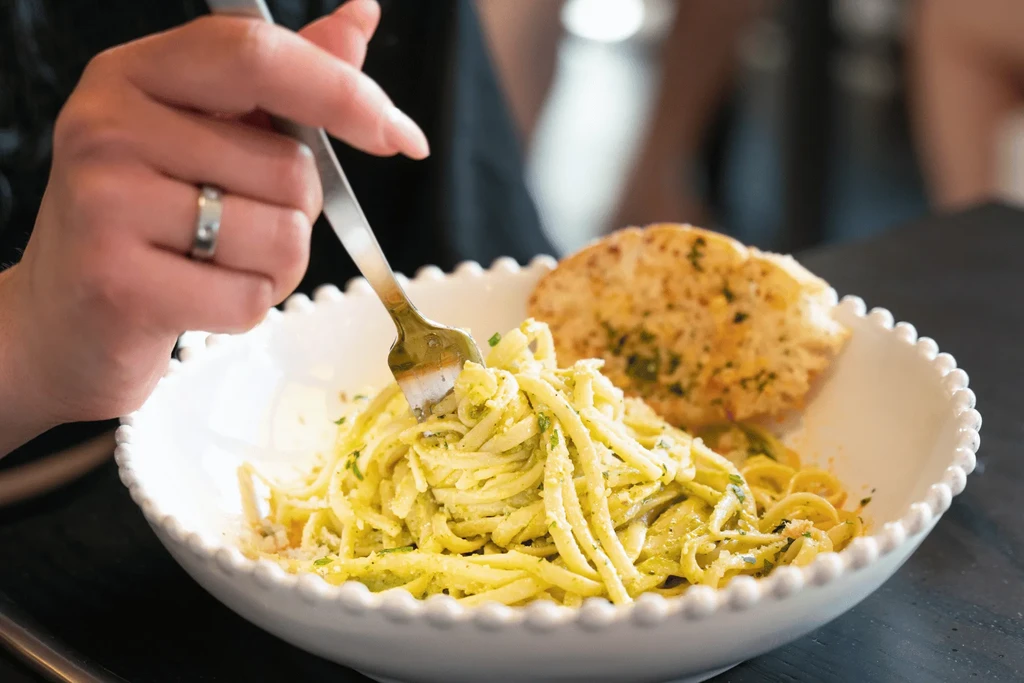 Hand twirling green pesto pasta with a fork and garlic bread.