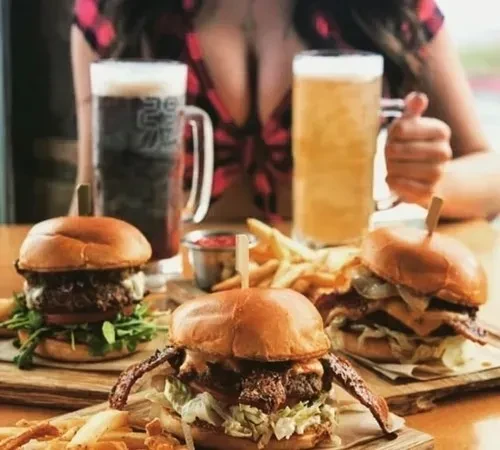Three gourmet burgers with fries, two beers, and a person's hand.