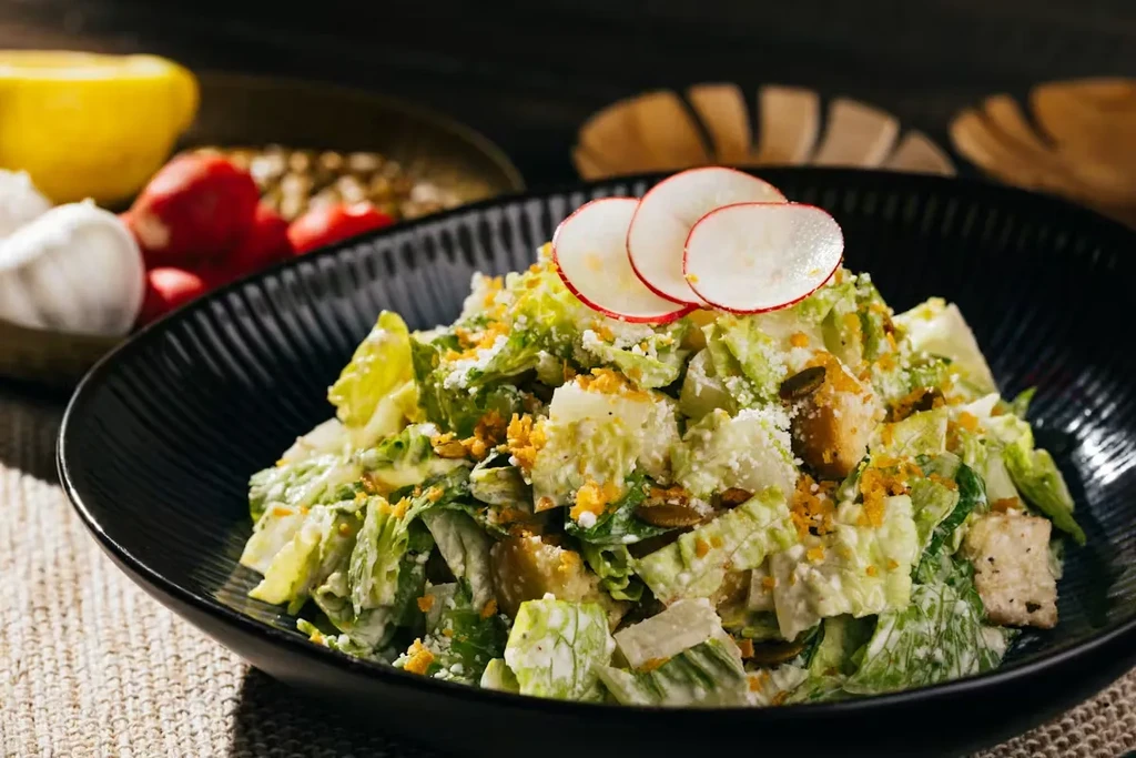 Caesar salad in dark ribbed bowl, with radishes, seeds, croutons.