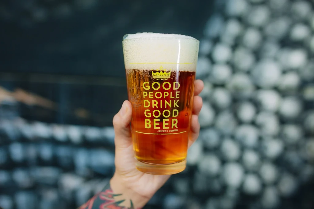 Tattooed hand holds amber beer glass with "GOOD PEOPLE DRINK GOOD BEER."