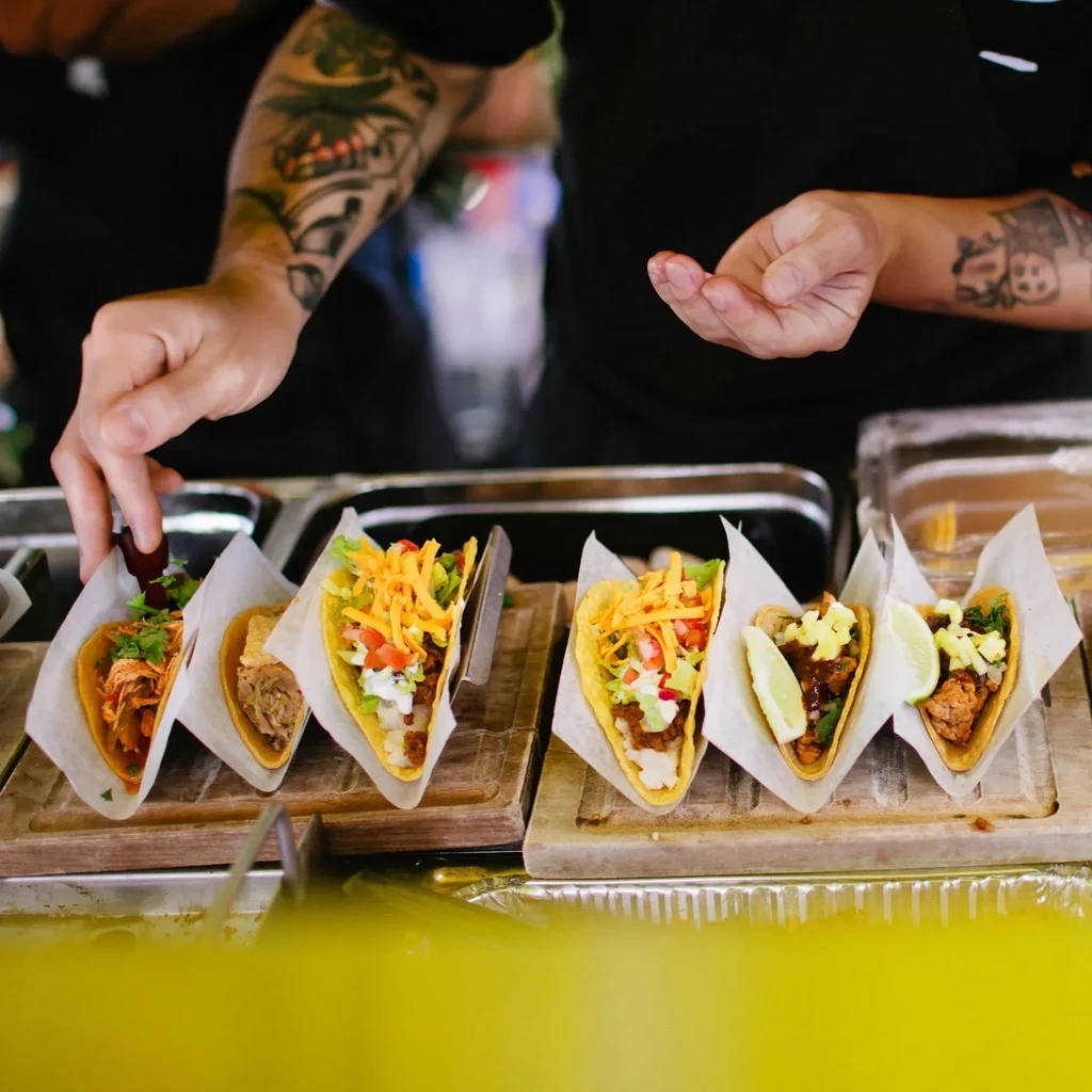Close-up of multiple tacos on wooden boards, touched by tattooed hands.