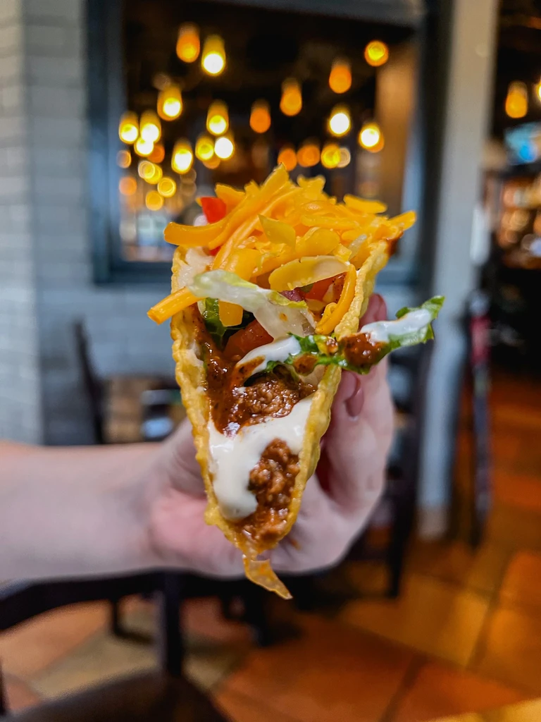 Hand holds a hard-shell taco filled with meat, cheese, lettuce, and sauce.