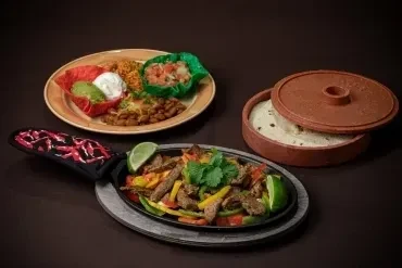 Sizzling fajitas in a skillet with tortillas and Mexican side dishes.