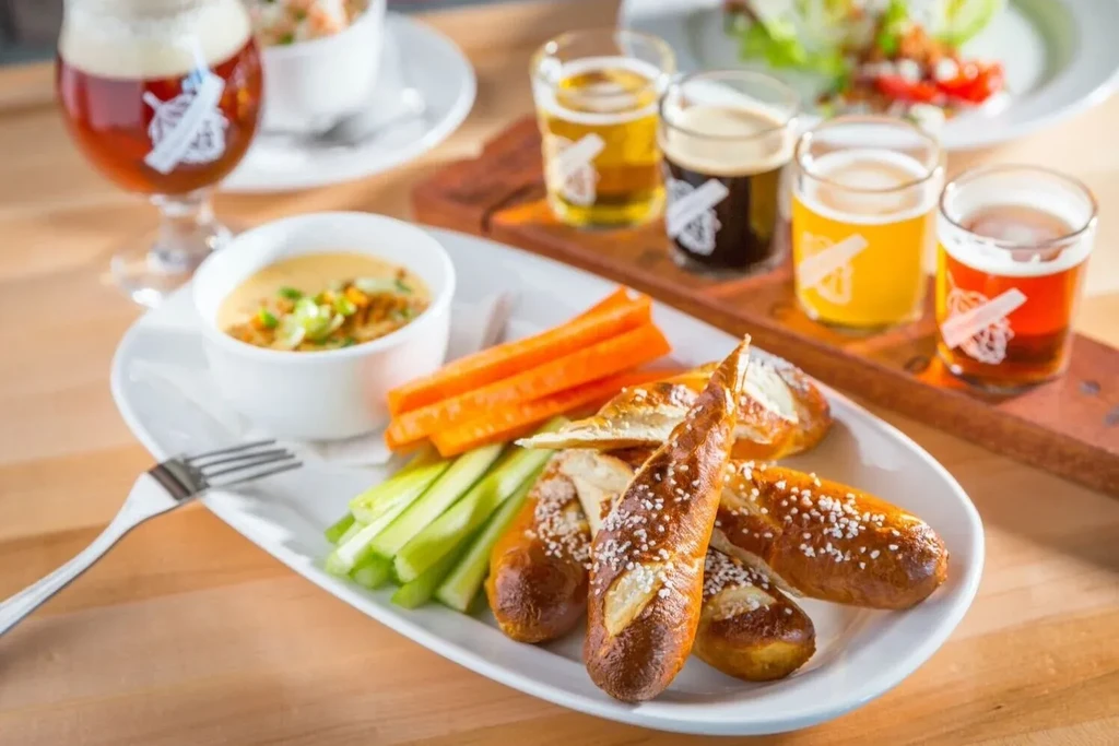 Pretzel sticks, vegetables, dip, and a beer flight on a wooden table.