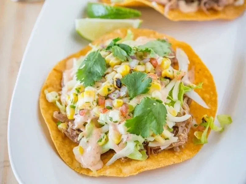 Tostada with shredded meat, lettuce, corn salsa, and drizzled sauce.