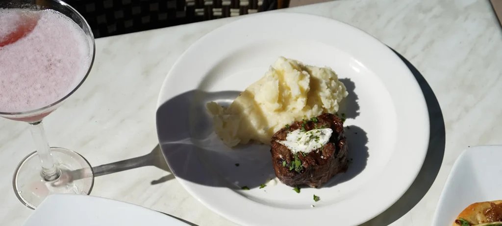 Grilled meat, mashed potatoes, pink drink on a bright stone table.