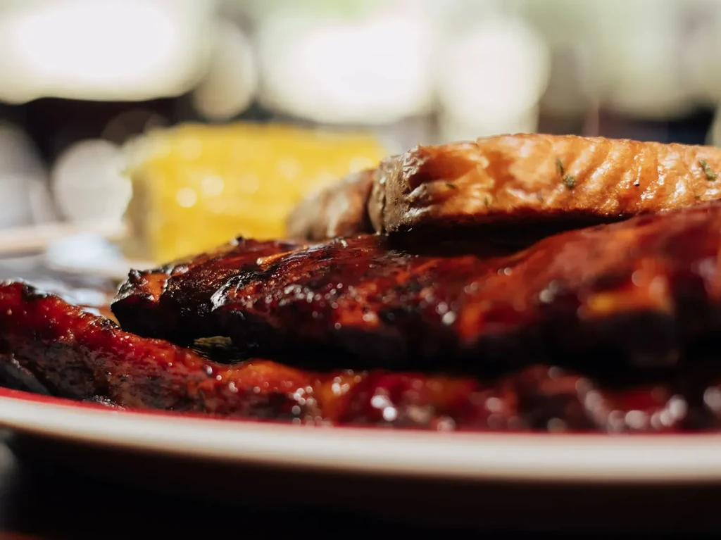 Glazed ribs with dark sauce, garnished food, and blurred corn.
