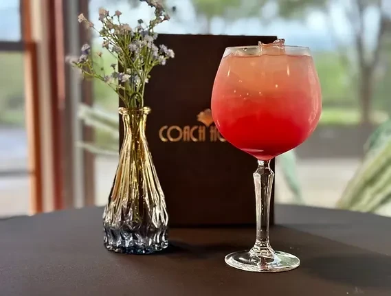 Red and pink drink in stemmed glass, next to a vase of flowers.