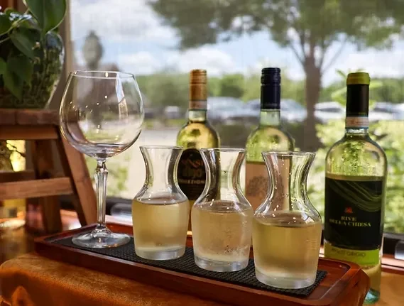 Pale wine carafes on wooden tray, bottles, by window.