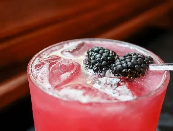 Pink iced drink in a glass, garnished with blackberries and a straw.