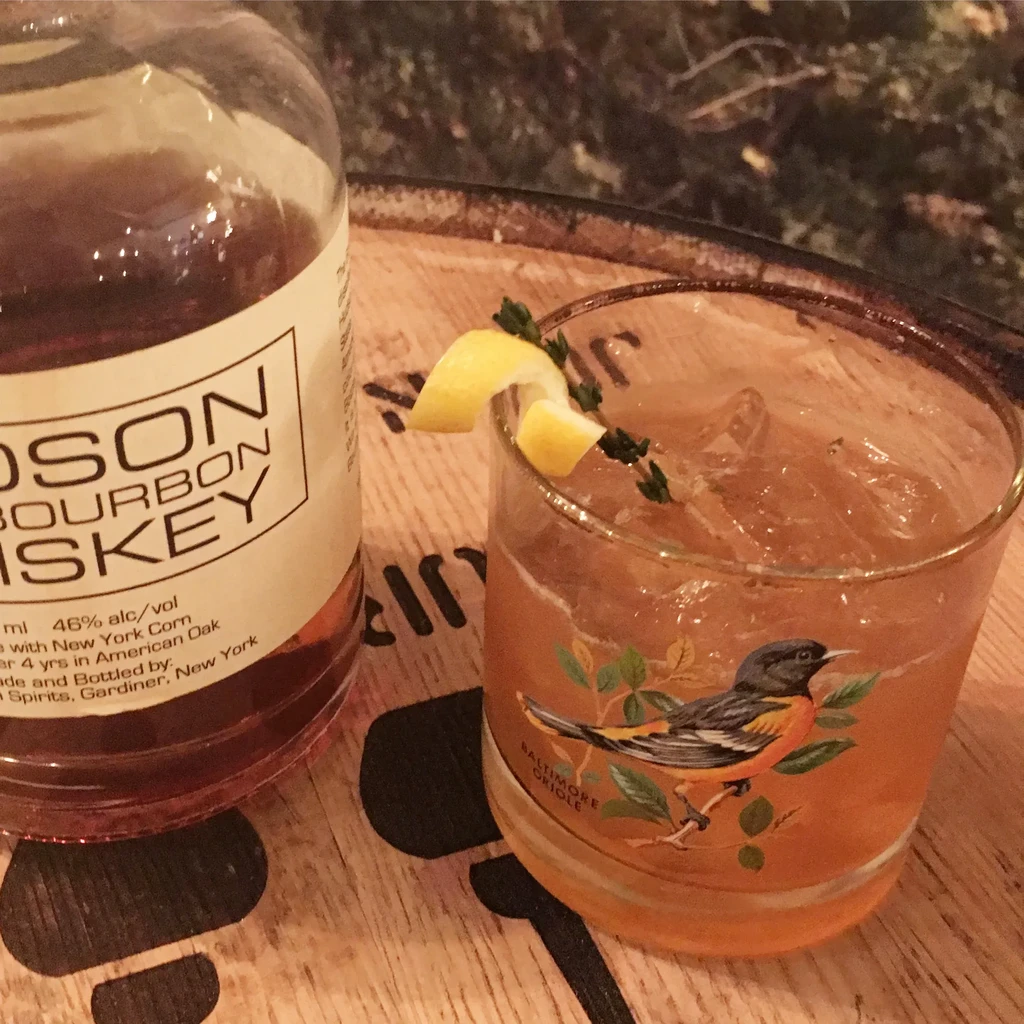 Hudson Bourbon whiskey bottle, Oriole-illustrated cocktail, on wood.