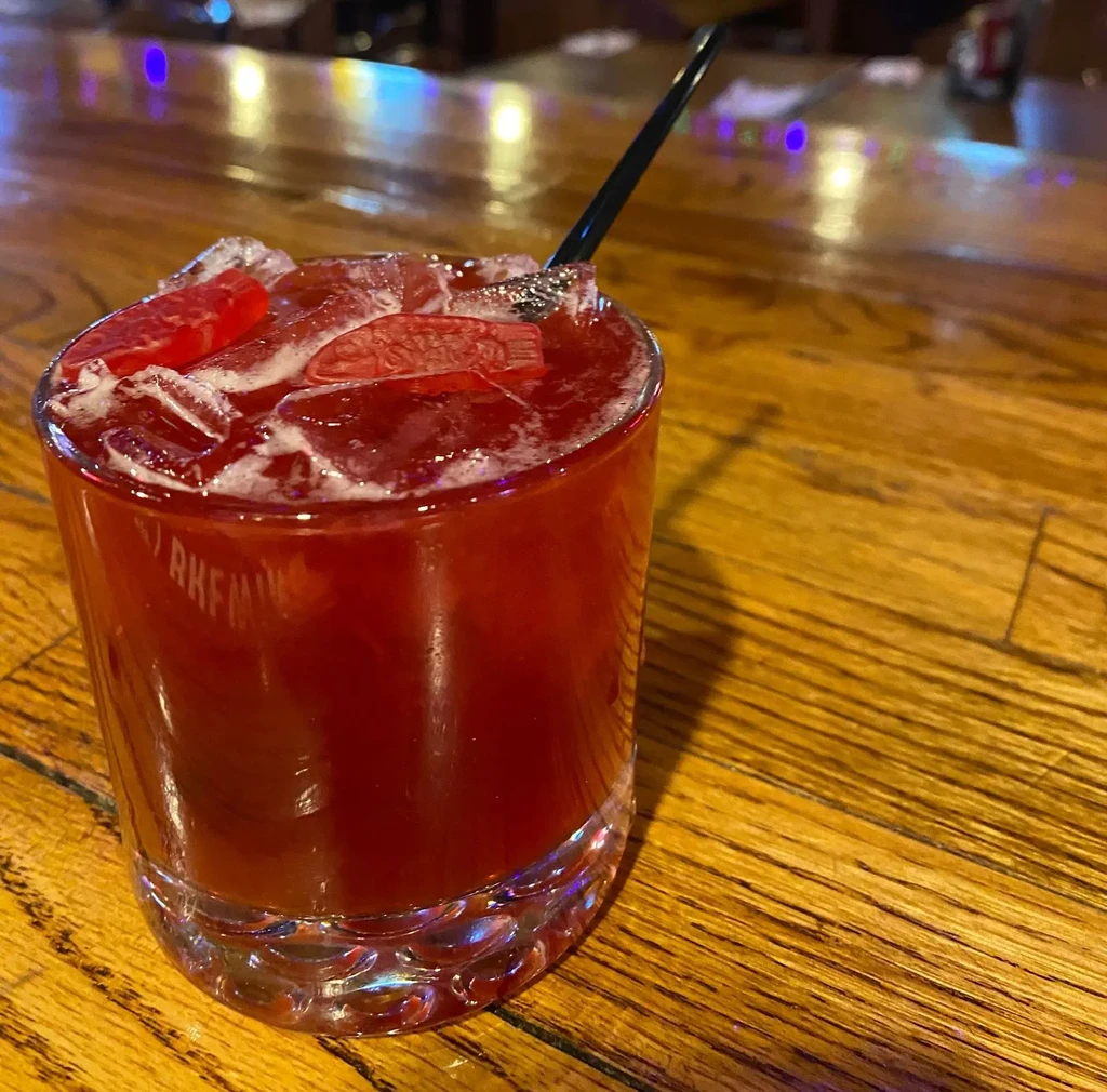 Red cocktail with foamy top, gummy candies, and a black straw.