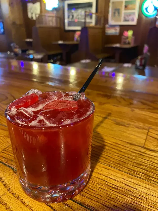 Red cocktail with fruit slices, ice, and straw on a bar.