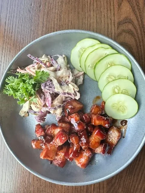 Glazed meat, colorful coleslaw, and fanned cucumber on a blue plate.