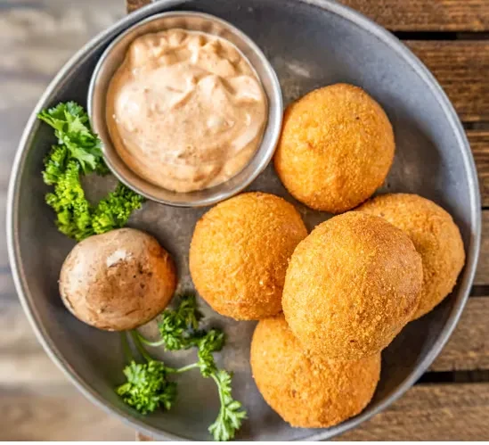 Six fried appetizers with dipping sauce, parsley, and mushroom on a rustic tray.