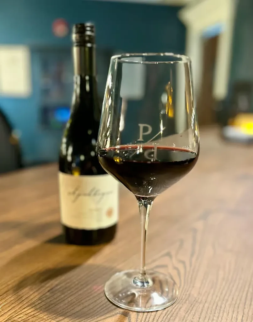 Etched 'P' wine glass with red wine and bottle on wooden table.