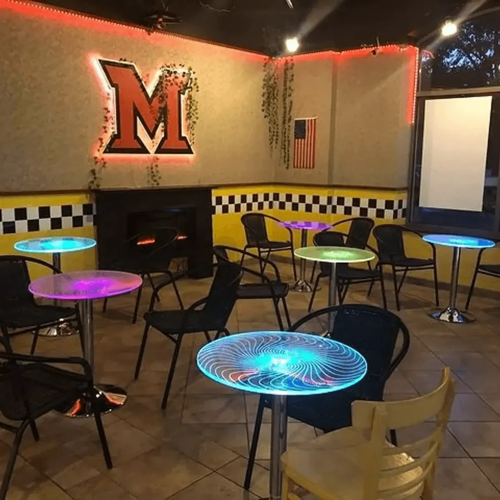 Modern restaurant with glowing tables, neon 'M' logo, and faux fireplace.