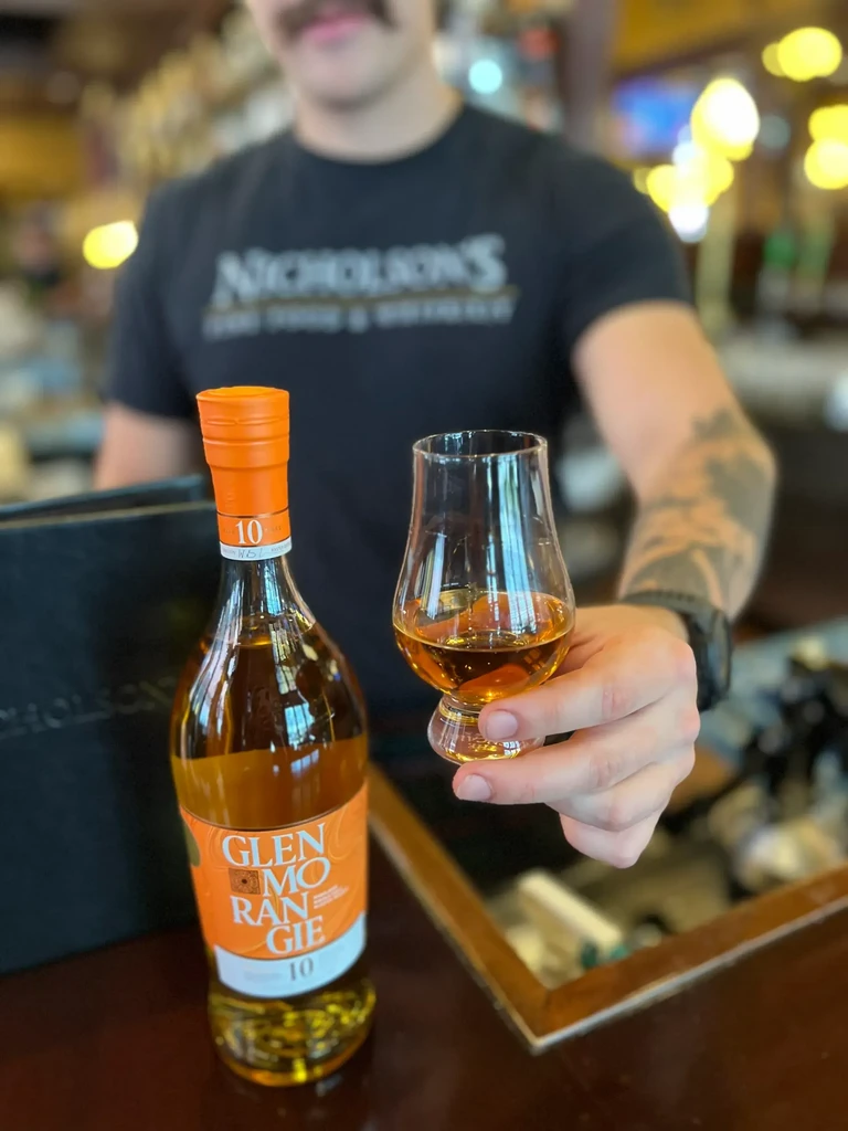 Glenmorangie 10-year scotch bottle, glass, blurred person with mustache at bar.