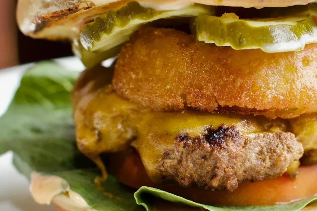 Close-up of a layered burger with onion ring, melted cheese, and pickles.