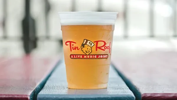 Plastic cup of cold beer with "Tin Roof" logo on wooden surface.