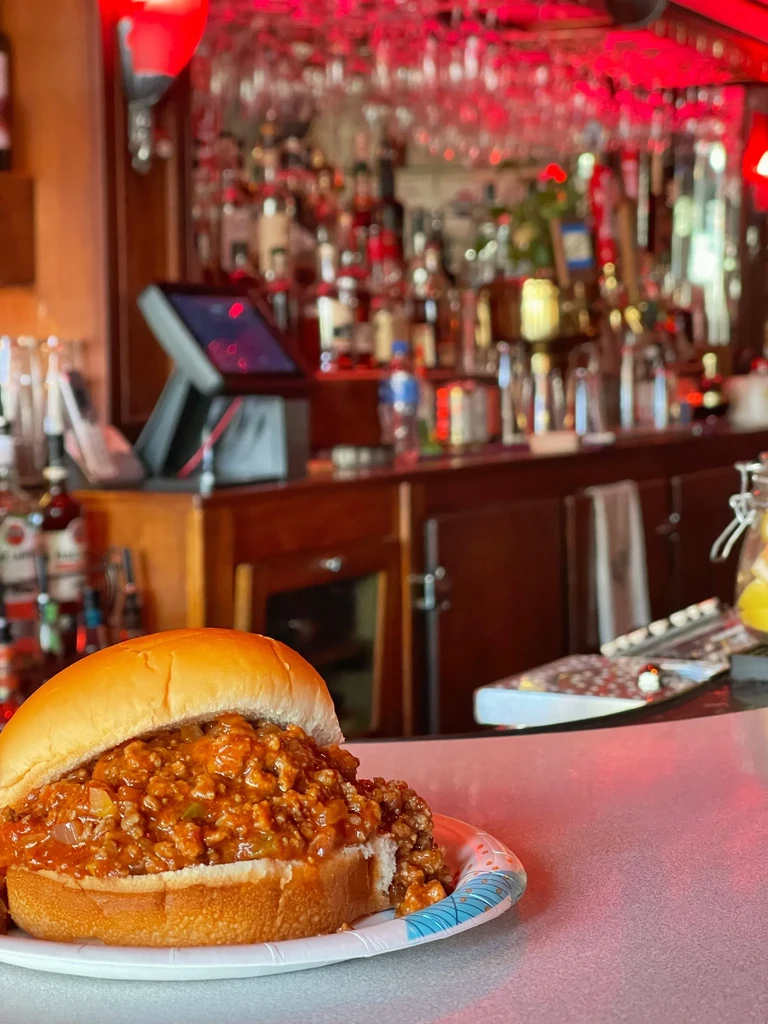 Sloppy joe on a paper plate with a blurred, red-lit bar background.