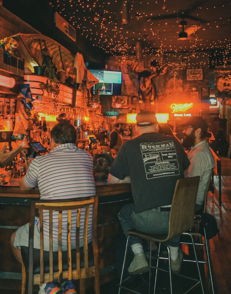 Cozy, rustic bar interior with string lights, patrons, and neon sign.
