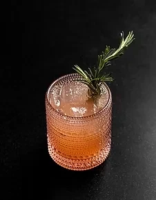 Rosemary garnished, light orange cocktail in a textured pink glass with ice.