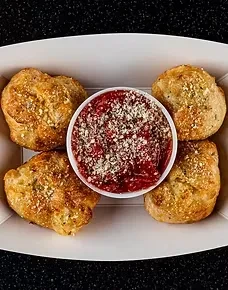 Four cheesy herb garlic knots with red dipping sauce in container.