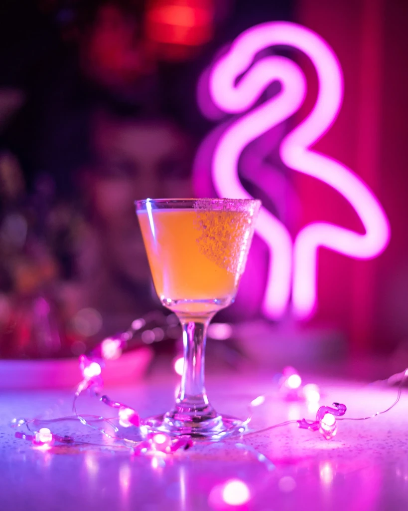 Yellow-orange cocktail on neon bar, with pink lights and flamingo.