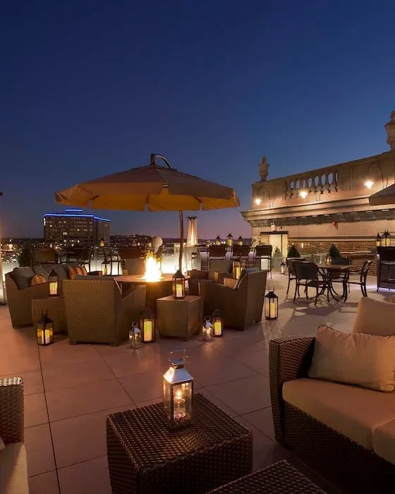 Rooftop lounge with fire pit, wicker furniture, lanterns, and city skyline.