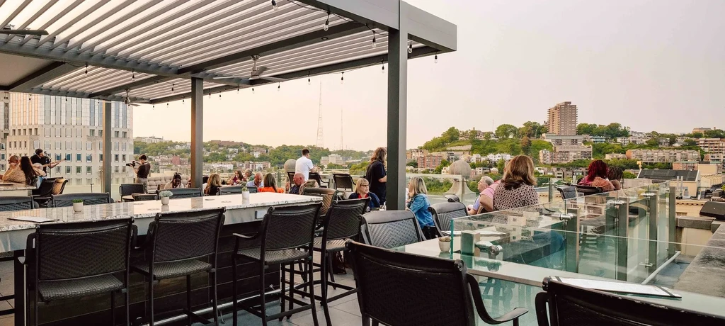 Rooftop restaurant with bar, pergola, people, and city skyline at dusk.