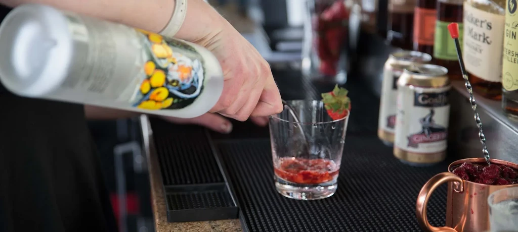 Hands pour clear liquid into a strawberry-garnished cocktail glass.