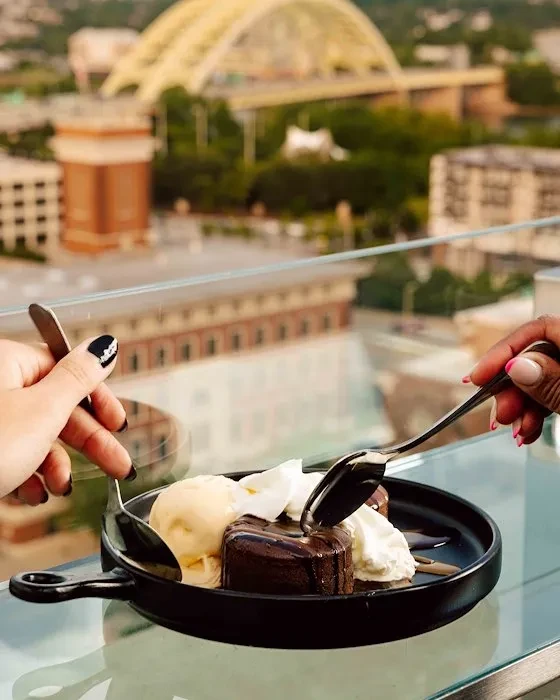 Chocolate lava cake with ice cream, two hands with spoons, blurred cityscape.