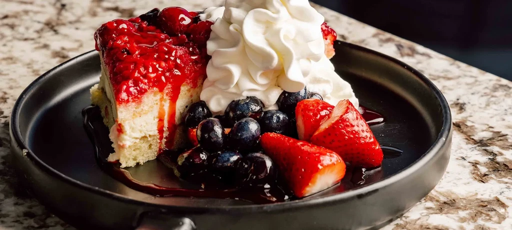 Cake slice with red berry sauce, whipped cream, blueberries, and strawberries.