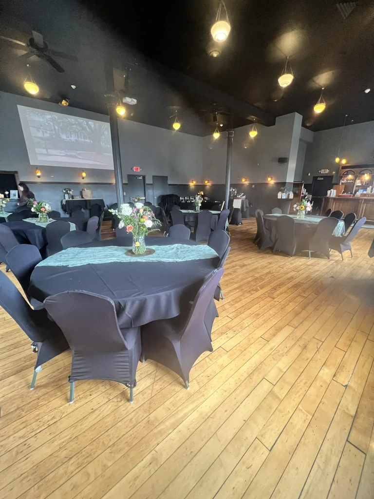 Spacious event venue with decorated tables, warm lighting, screen, and bar.