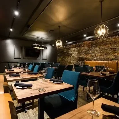 Restaurant interior with wooden tables, teal chairs, stone wall, warm lighting.