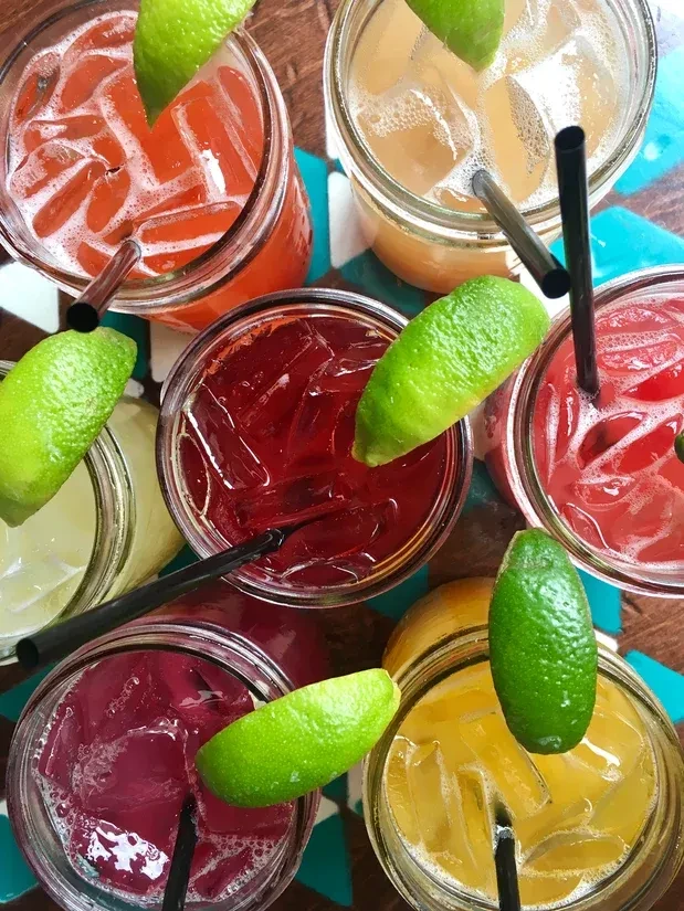 Six colorful iced drinks in jars with lime wedges and straws.