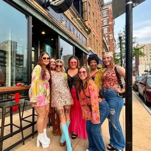 Seven smiling women in retro outfits posing outside a Pinball arcade.