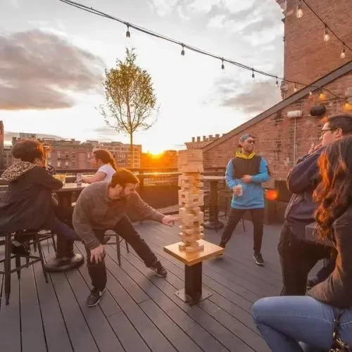Young adults play giant Jenga at a lively rooftop bar at sunset.