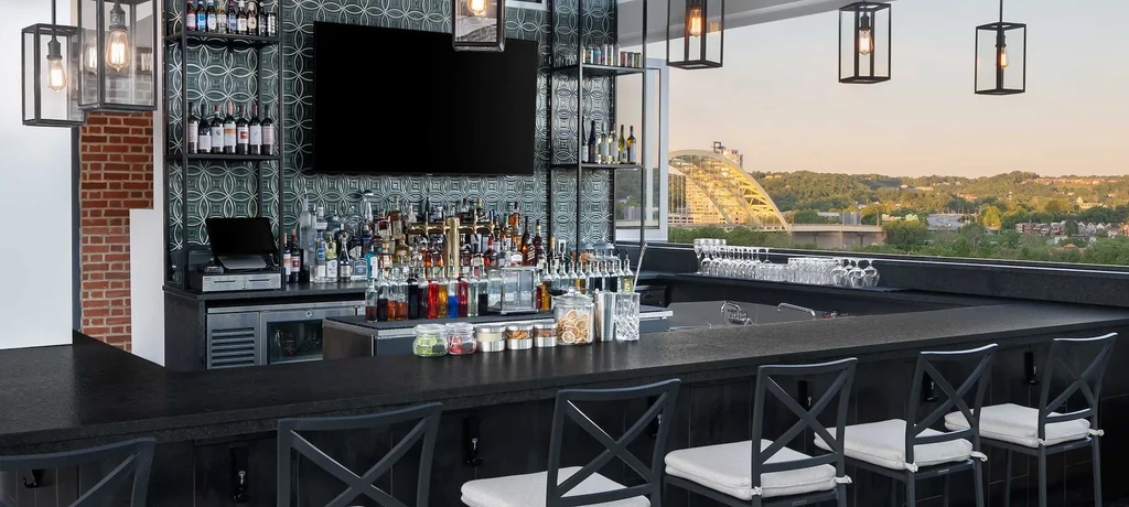Well-stocked outdoor bar overlooks city, green hills, and bridge at sunset.