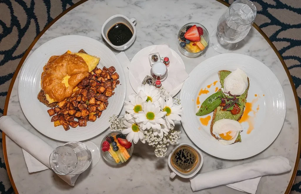 Overhead view: Brunch spread with croissant, avocado toast, eggs, coffee, flowers.