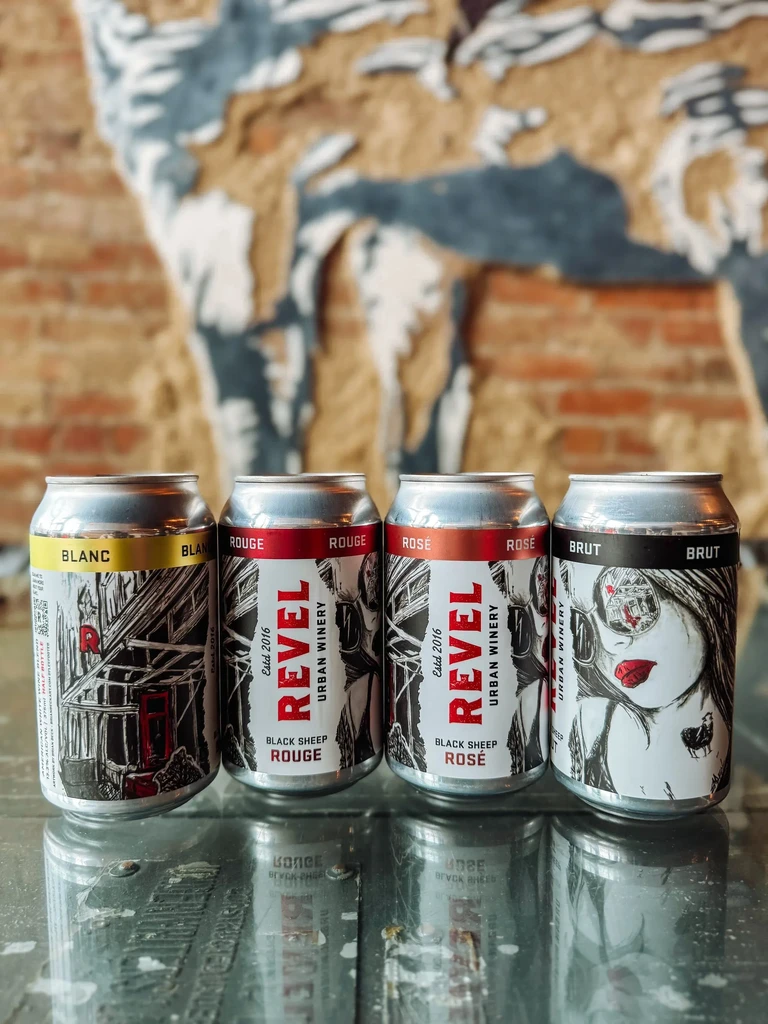 Four Revel Urban Winery cans in a row on a reflective surface.