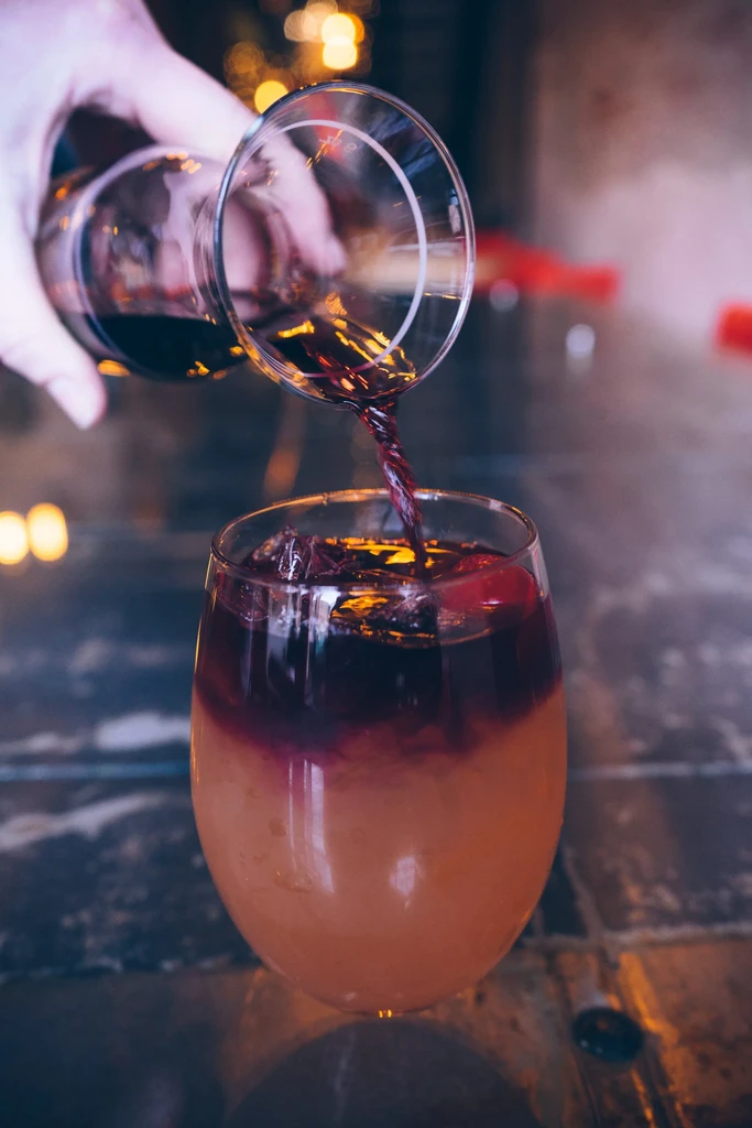 Hand pouring dark liquid into a two-toned drink with ice.
